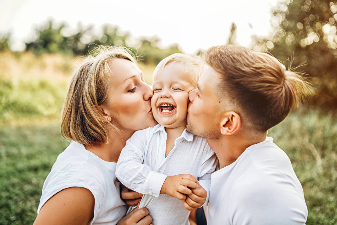 Ein blonder Junge wird von seinen Eltern gehalten und bekommt von Vater und Mutter einen Kuss auf je eine Wange. | © Adobestock