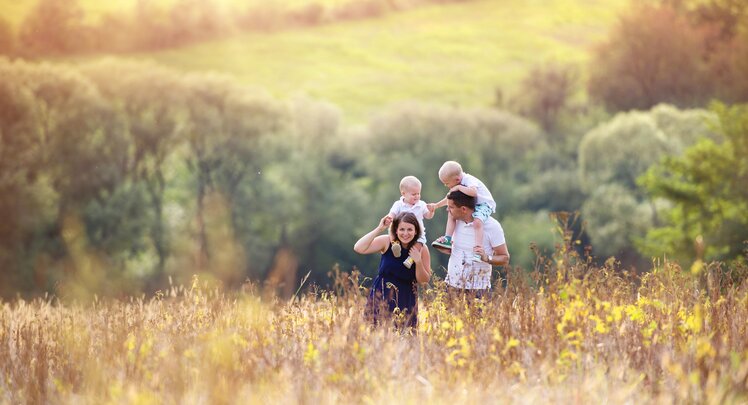 Eine Familie mit 2 Kindern spaziert in der Abendsonne durch ein trockenes Blumenfeld. Die Eltern haben beide je ein Kind auf den Schultern.