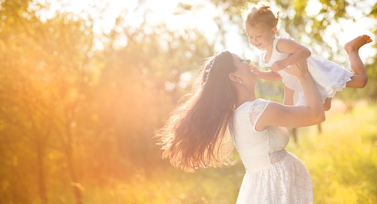 Eine schwangere Frau in weissem Kleid hebt ihre kleine Tochter in weissem Kleid in die Luft. Sie lachen einander an. | © Adobestock