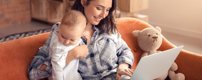 Mutter sitzt mit Baby auf dem Arm auf dem Sofa an einem Laptop | © Adobe Stock