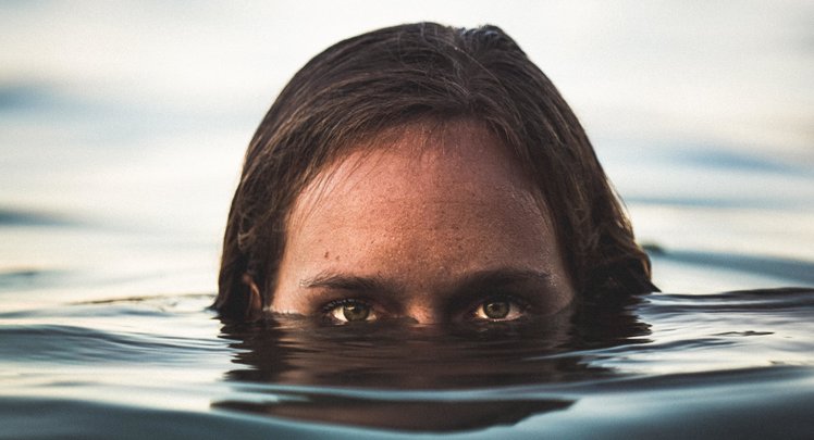 Eine braunhaarige Frau mit grünen Augen taucht aus dem Wasser auf. Ihre Augen sind knapp über der Wasserlinie.
