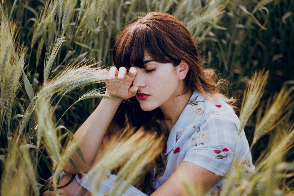 Eine Frau mit braunen Augen sitzt in einem trockenen Weizenfeld. Sie ist müde und hält eine Hand aufs Auge. | © Hazzel Silva - Unsplash