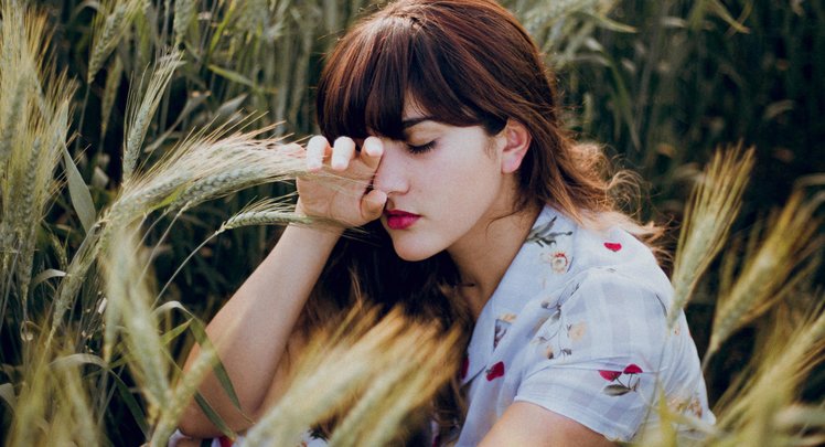 Eine Frau mit braunen Augen sitzt in einem trockenen Weizenfeld. Sie ist müde und hält eine Hand aufs Auge. | © Hazzel Silva - Unsplash