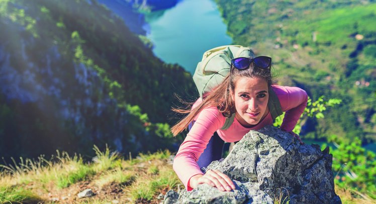 Eine Frau klettert in den Bergen einen Fels hoch und schaut von unten hoch in die Kamera. | © Adobestock