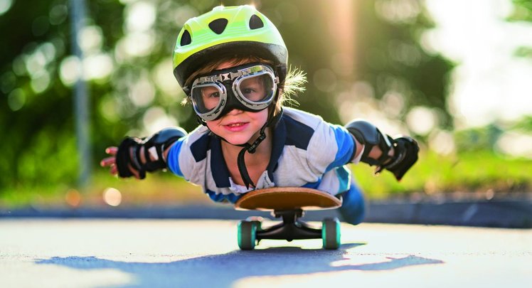 Ein Junge liegt auf einem fahrenden Skateboard. Er trägt einen Helm, Ellbogen- & Handschoner und eine Fliegerbrille. | © iStock