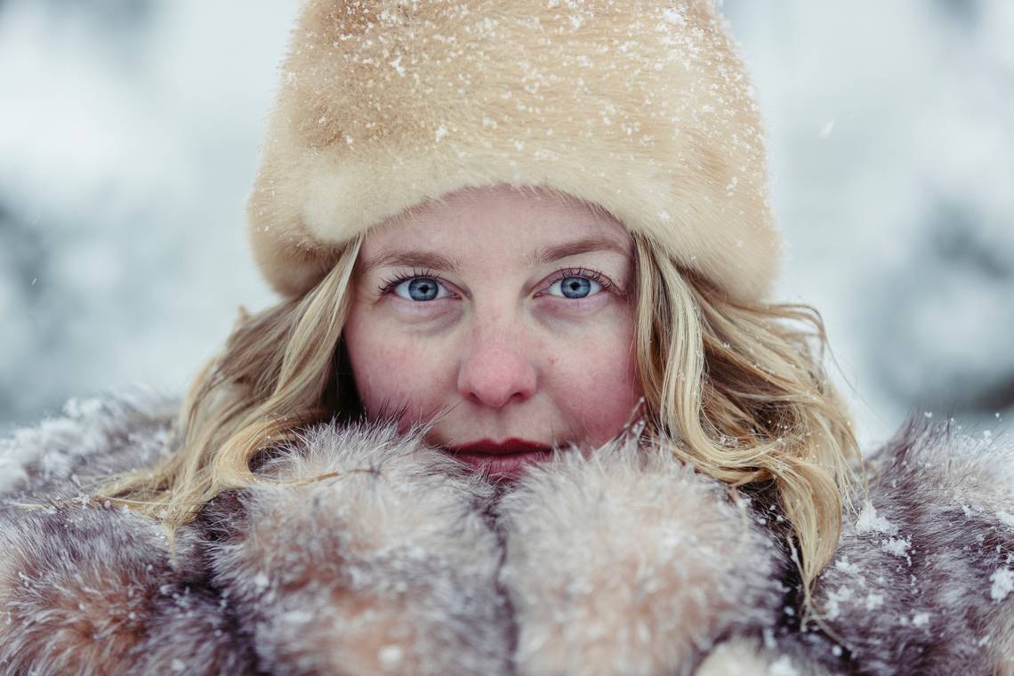Frau mit blonden Haaren, blauen Augen und einer Fellmütze hält sich die Hände in Handschuhen vor das Gesicht in Winterlandschaft. | © Martin Reisch_via Unsplash