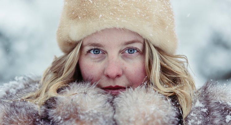 Frau mit blonden Haaren, blauen Augen und einer Fellmütze hält sich die Hände in Handschuhen vor das Gesicht in Winterlandschaft. | © Martin Reisch_via Unsplash