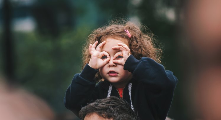 Ein rothaariges Mädchen sitzt auf den Schultern eines Mannes und schaut durch ihre Brille, die sie mit Daumen und Zeigefinger formt. | © Edi Libedinsky via Unsplash