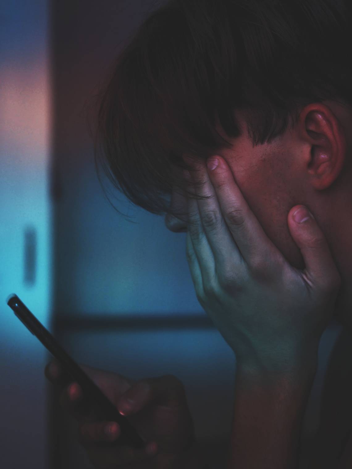 Ein junger Mann hält sein Mobile Phone in dunkelm Licht vor dem Gesicht und reibt sich mit der anderen Hand die Augen. | © Adrian Swancar via Unsplash