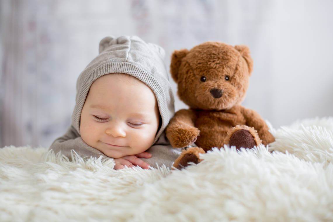 Schlafendes Baby, das ein Lächeln auf dem Gesicht hat. Daneben sitzt ein Teddybär.