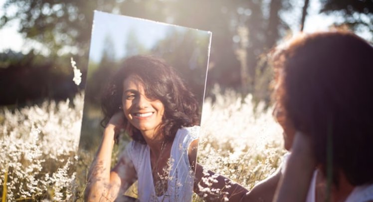 Frau sitzt in Wiese und lächelt in einen Spiegel, den sie hält. | © Caroline Veronez via Unsplash
