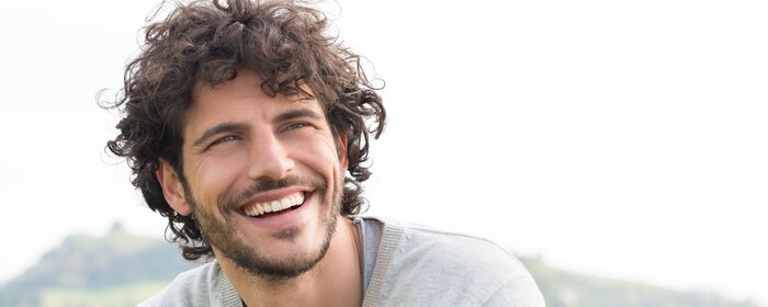 Lachender Mann mit dunkelbraunen Locken, 3-Tage-Bart und hellgrauem Pullover. | © Shutterstock