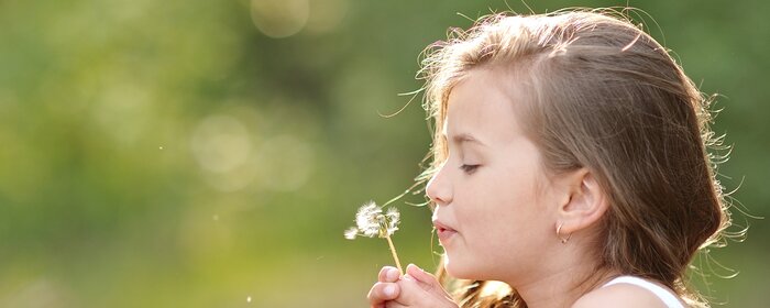 Mädchen mit dunkelblonden Haaren hält eine Pusteblume in der Hand und pustet. | © Shutterstock