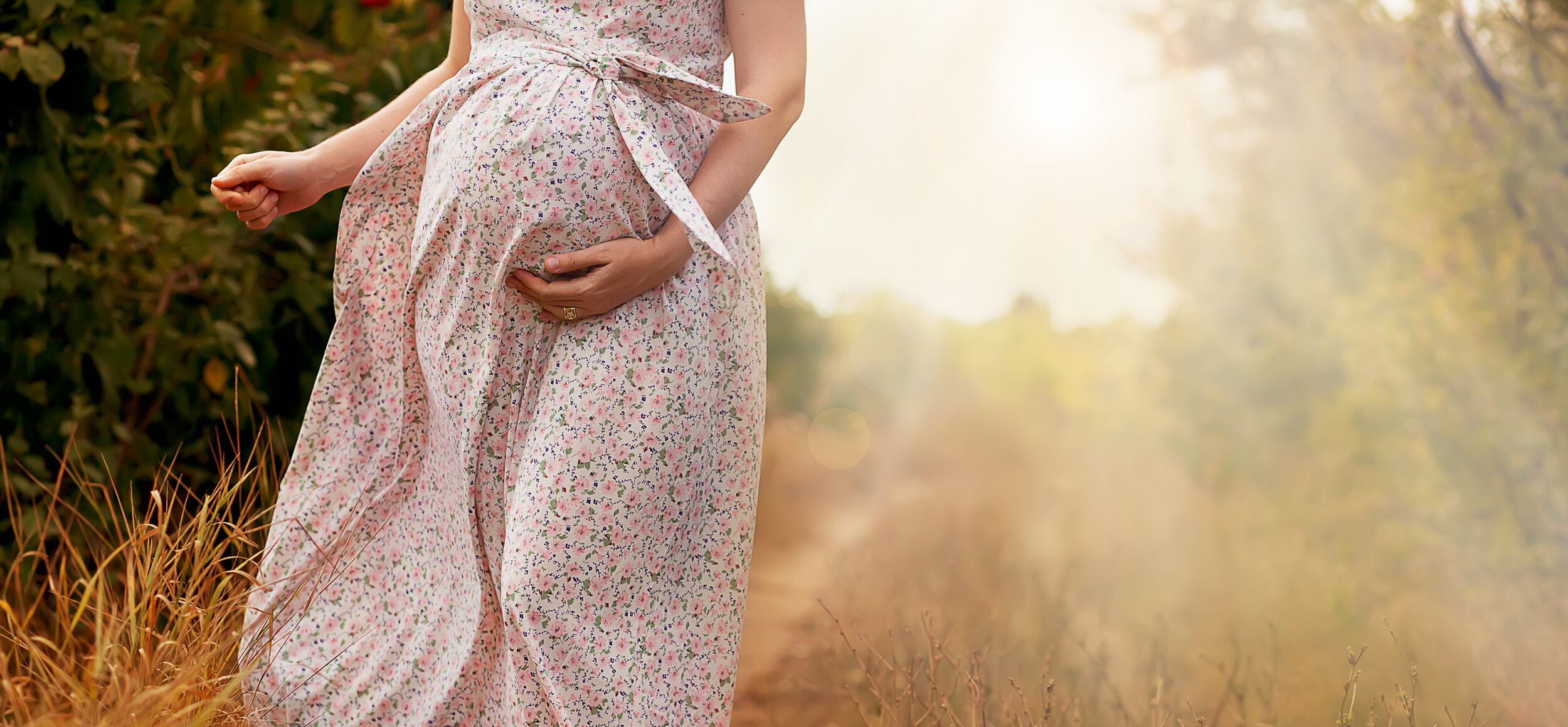 Schwangere Frau steht barfuss auf einem Waldweg in einem geblümten Kleid.